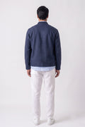A man viewed from behind wearing a navy blue linen zip-up jacket over a light blue shirt, paired with off-white linen trousers and white sneakers, against a plain light gray studio background.