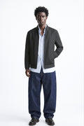 A man modeling a dark gray zip-up linen bomber jacket over a white button-down shirt, paired with loose dark blue jeans and black leather shoes, against a plain white studio background.