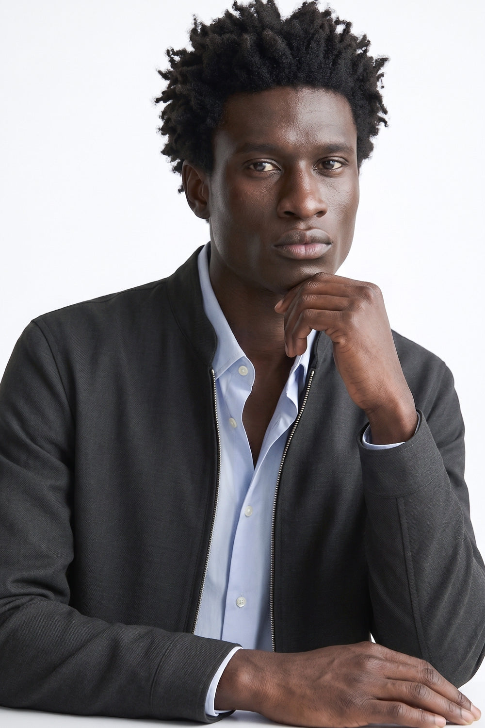 A man with short dark curly hair wearing a dark gray zip-up linen jacket over a light blue button-down shirt, posing against a plain white background.