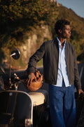 A man stands outdoors beside a scooter, wearing a dark gray linen bomber jacket over a light blue button-up shirt and blue jeans, holding a brown leather helmet. Sunlight casts a warm golden hour glow across the scene with a blurred hillside background.