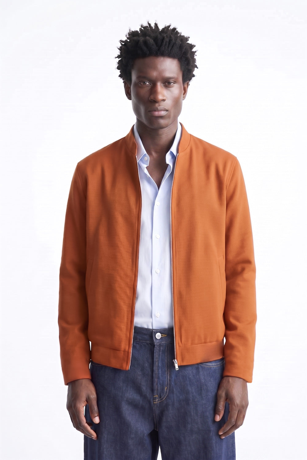 A man wearing a rust-orange zip-up linen bomber jacket over a white button-down shirt and dark blue jeans, standing against a plain white studio background.