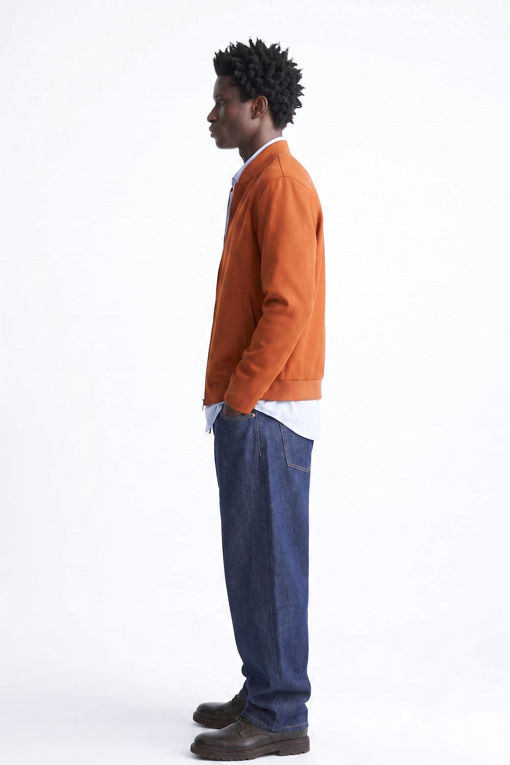 A man in profile wearing an orange linen jacket over a white shirt, blue jeans, and brown leather shoes, standing against a plain white studio background.