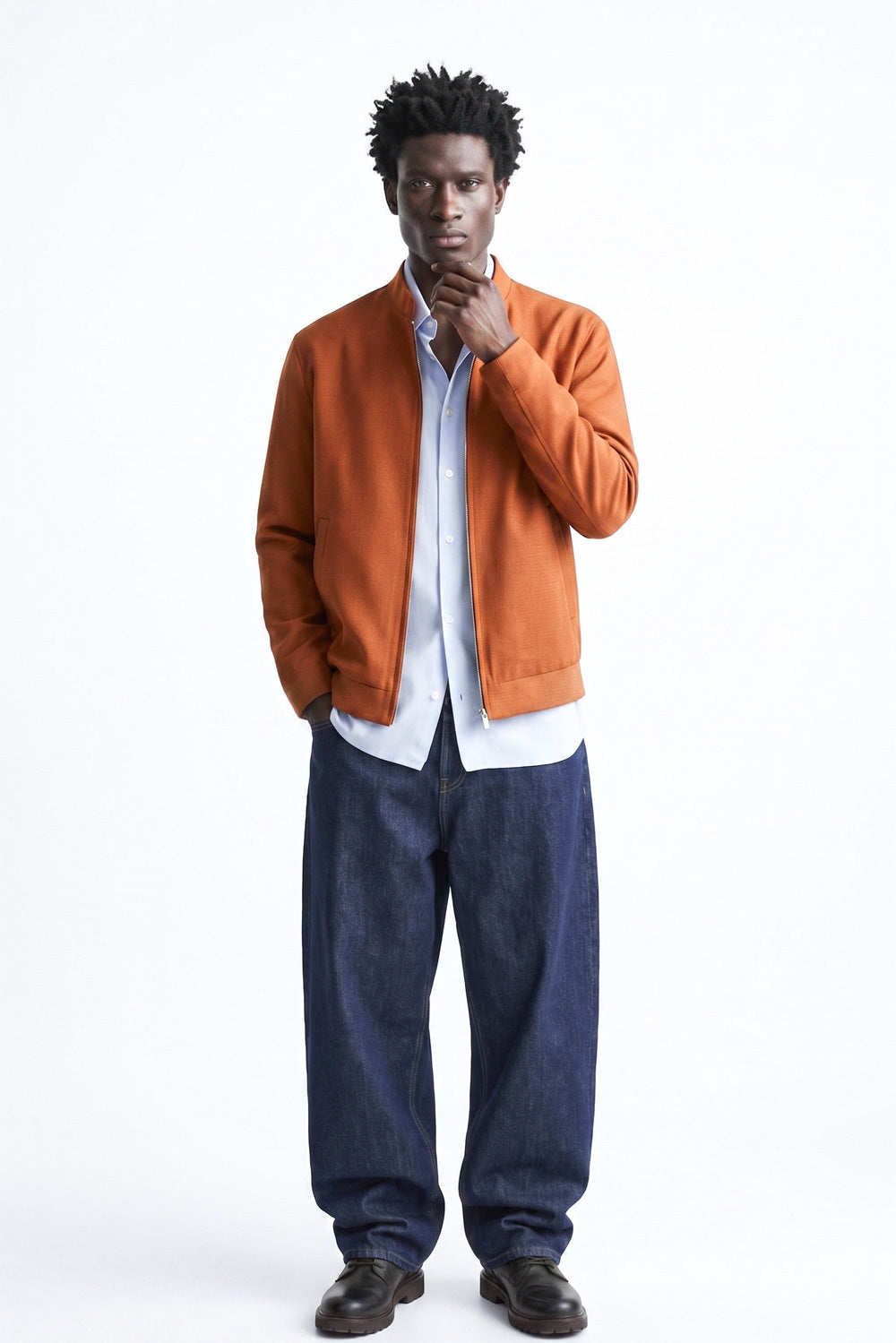 A man modeling a rust-orange linen jacket over a white button-down shirt and loose dark blue jeans, standing against a plain white studio background.