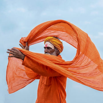 A Indian Sadhu in an orange robe and yellow turban holds a large flowing orange fabric against a pale blue sky.