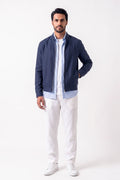A man wearing a navy blue zip-up linen jacket over a light blue button-down shirt and white pants, standing against a plain white studio background.