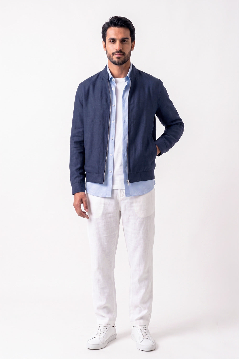 A man wearing a navy blue zip-up linen jacket over a light blue button-down shirt and white pants, standing against a plain white studio background.