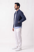 A man wearing a navy blue zip-up linen jacket over a light blue shirt, paired with white linen trousers and white sneakers, standing in profile against a plain white studio background.