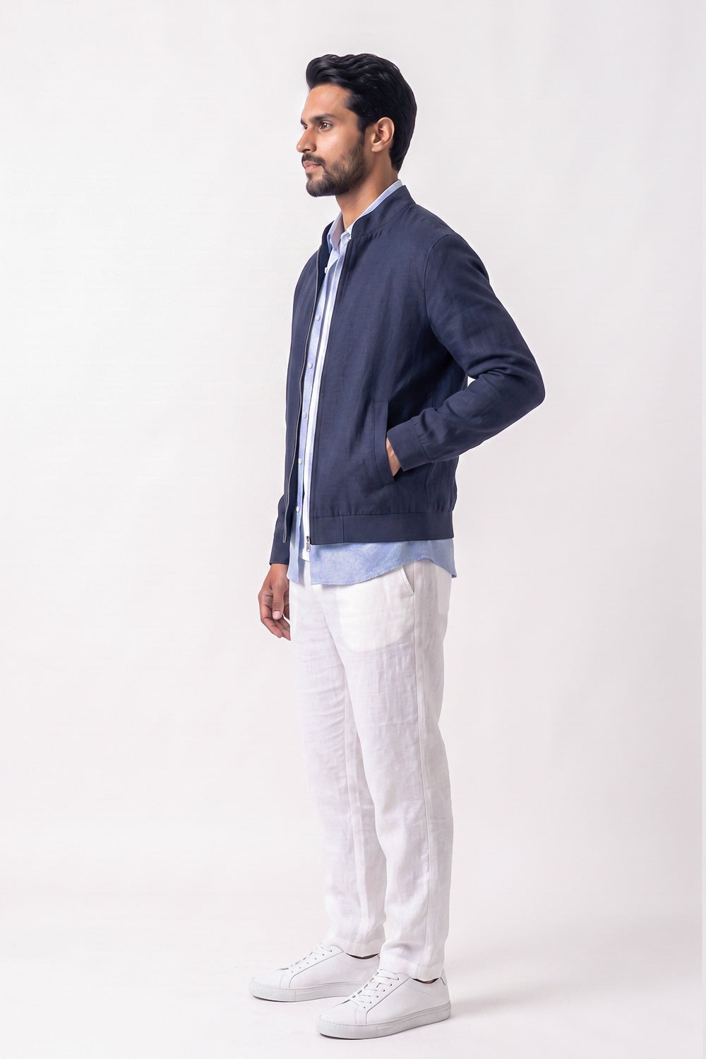 A man wearing a navy blue zip-up linen jacket over a light blue shirt, paired with white linen trousers and white sneakers, standing in profile against a plain white studio background.