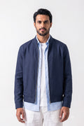 A man wearing a navy blue zip-up linen jacket over a light blue button-down shirt and white pants, standing against a plain white studio background.