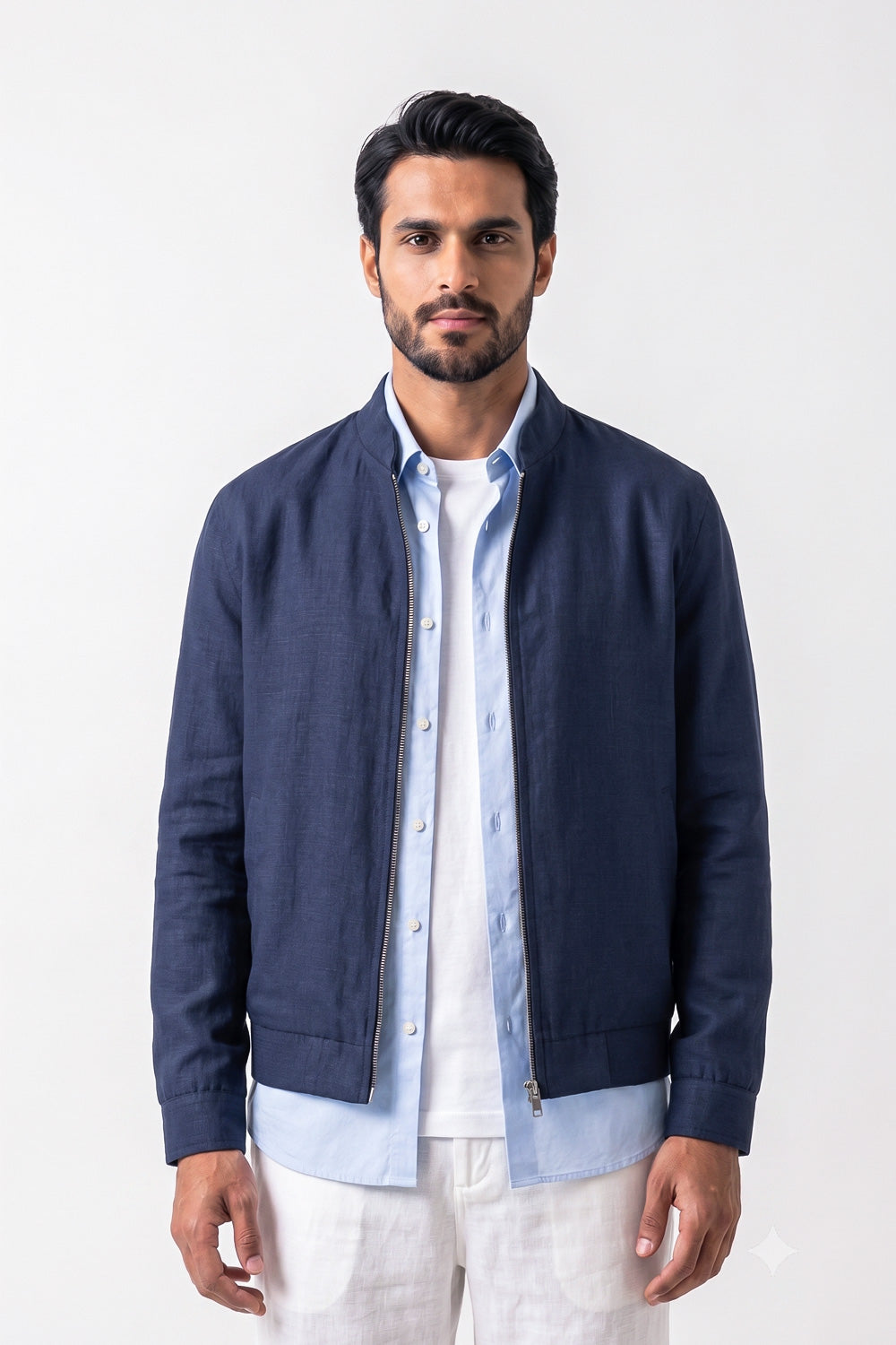 A man wearing a navy blue zip-up linen jacket over a light blue button-down shirt and white pants, standing against a plain white studio background.