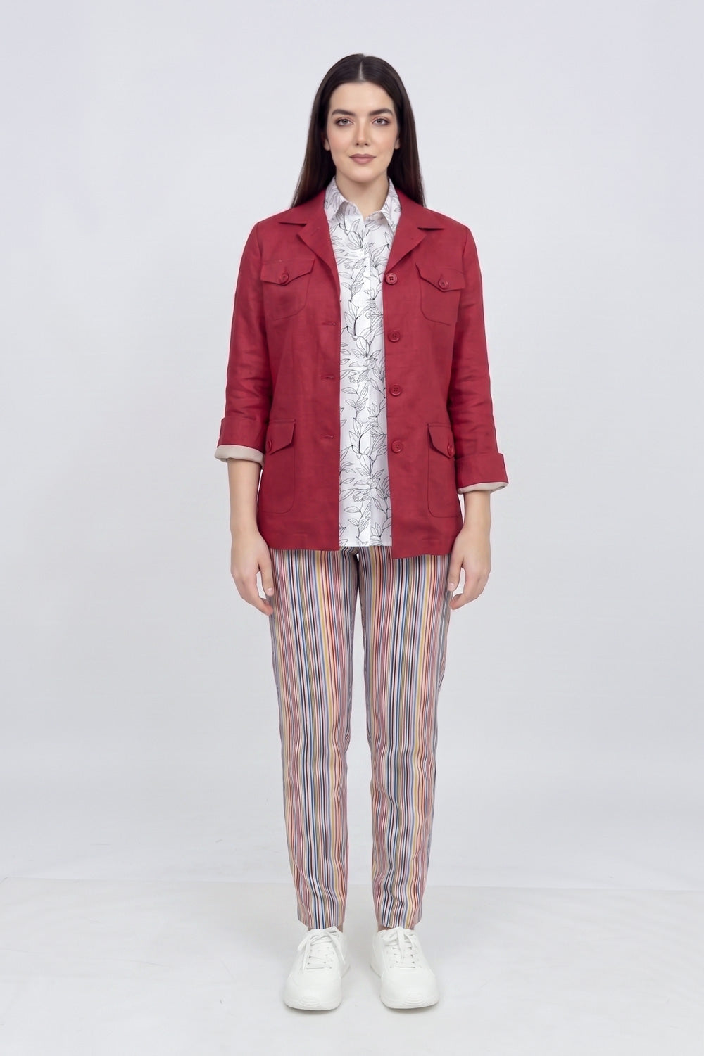 A woman stands against a light gray studio background wearing a red cropped linen jacket over a white patterned shirt, with vertically striped pants in pink, blue, and orange, and white sneakers.