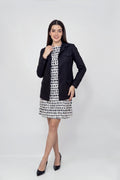 A woman modeling a black tailored linen jacket over a black-and-white patterned dress, paired with black pointed heels, against a light gray studio background.