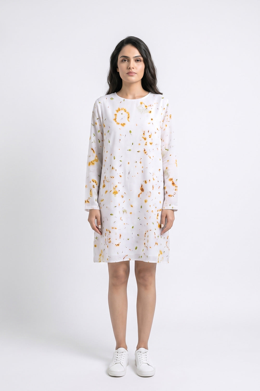 A woman modeling a white long-sleeve shift linen dress with a yellow and orange eco-print, paired with white sneakers, against a light gray studio background.