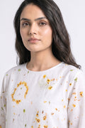 A woman wearing a white long-sleeve linen top with a yellow and greeneco-print floral pattern, shown in a close-up portrait against a light gray background.