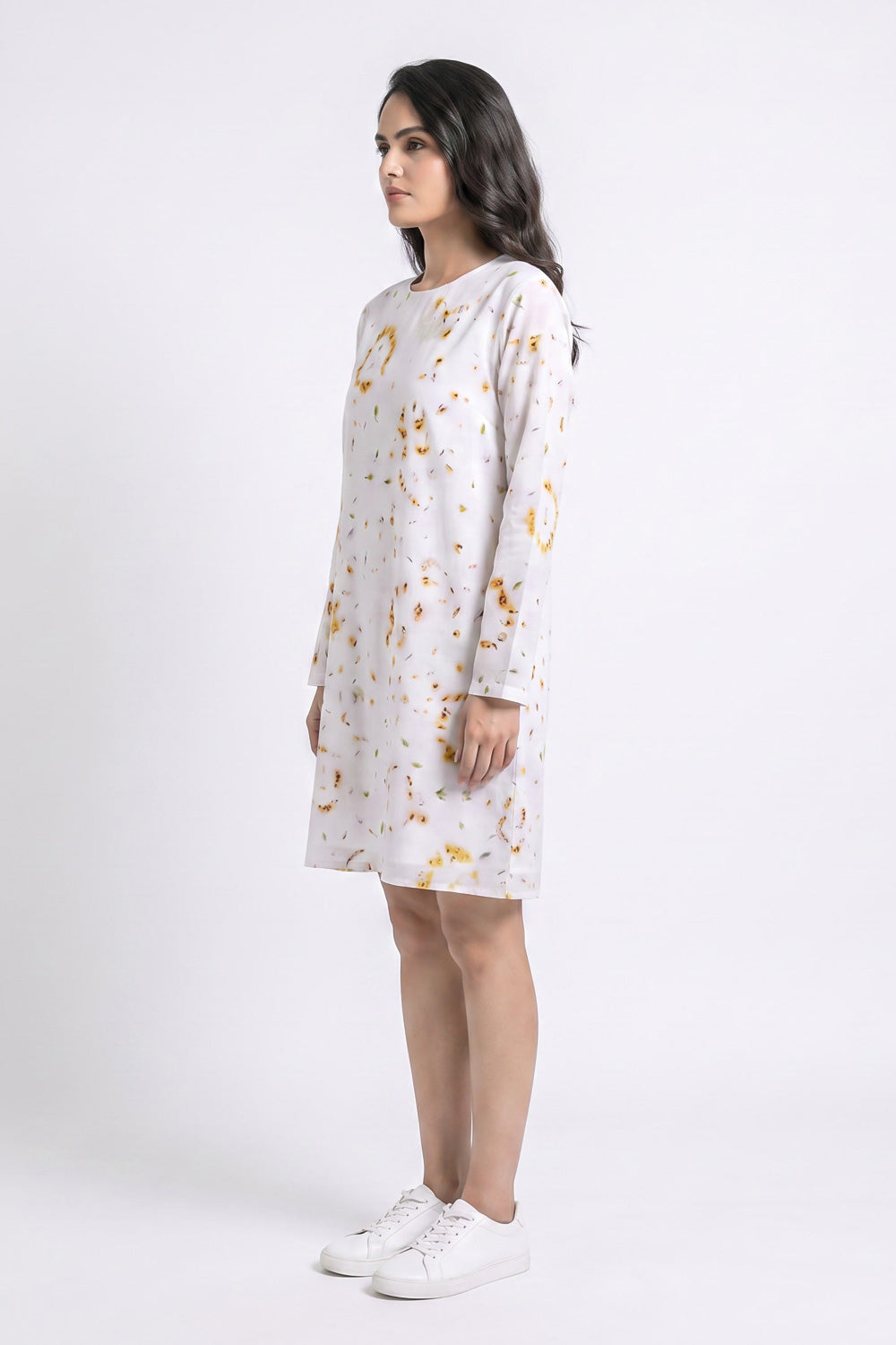 A woman modeling a long-sleeve white linen dress with a gold eco-print, paired with white sneakers, photographed against a plain white studio background.