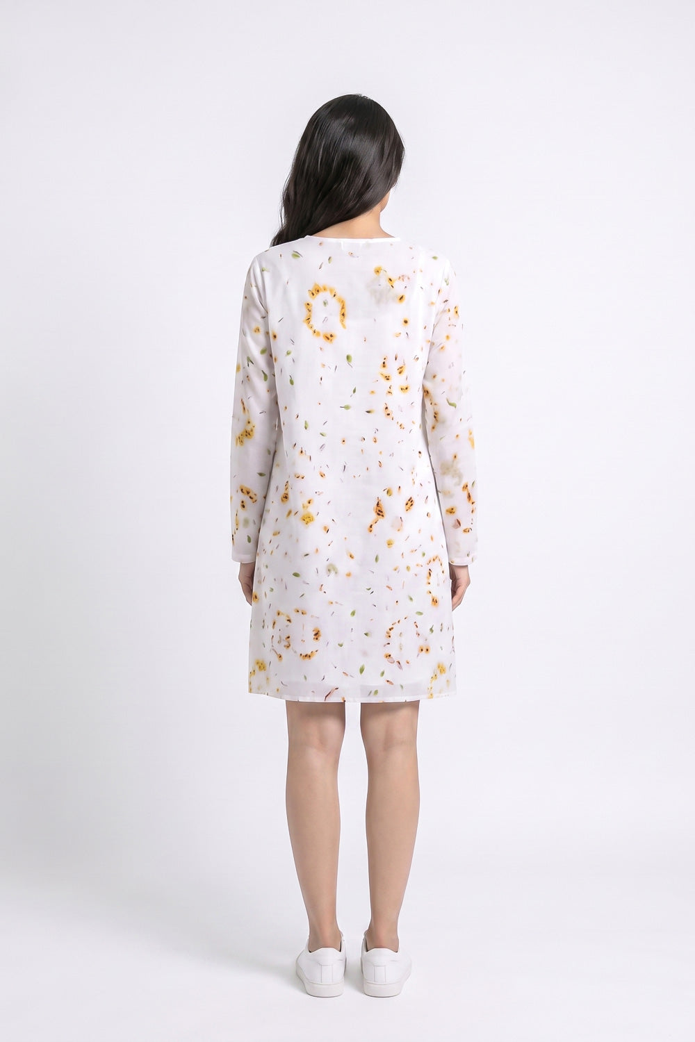 A woman viewed from the back wearing a long-sleeve linen white dress with a scattered orange and yellow eco-print, paired with white slip-on shoes, against a plain white studio background.