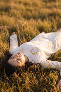 A woman lying on her back in a grassy field during golden hour, wearing a white long-sleeve linen top with yellow eco-print style circular patterns.