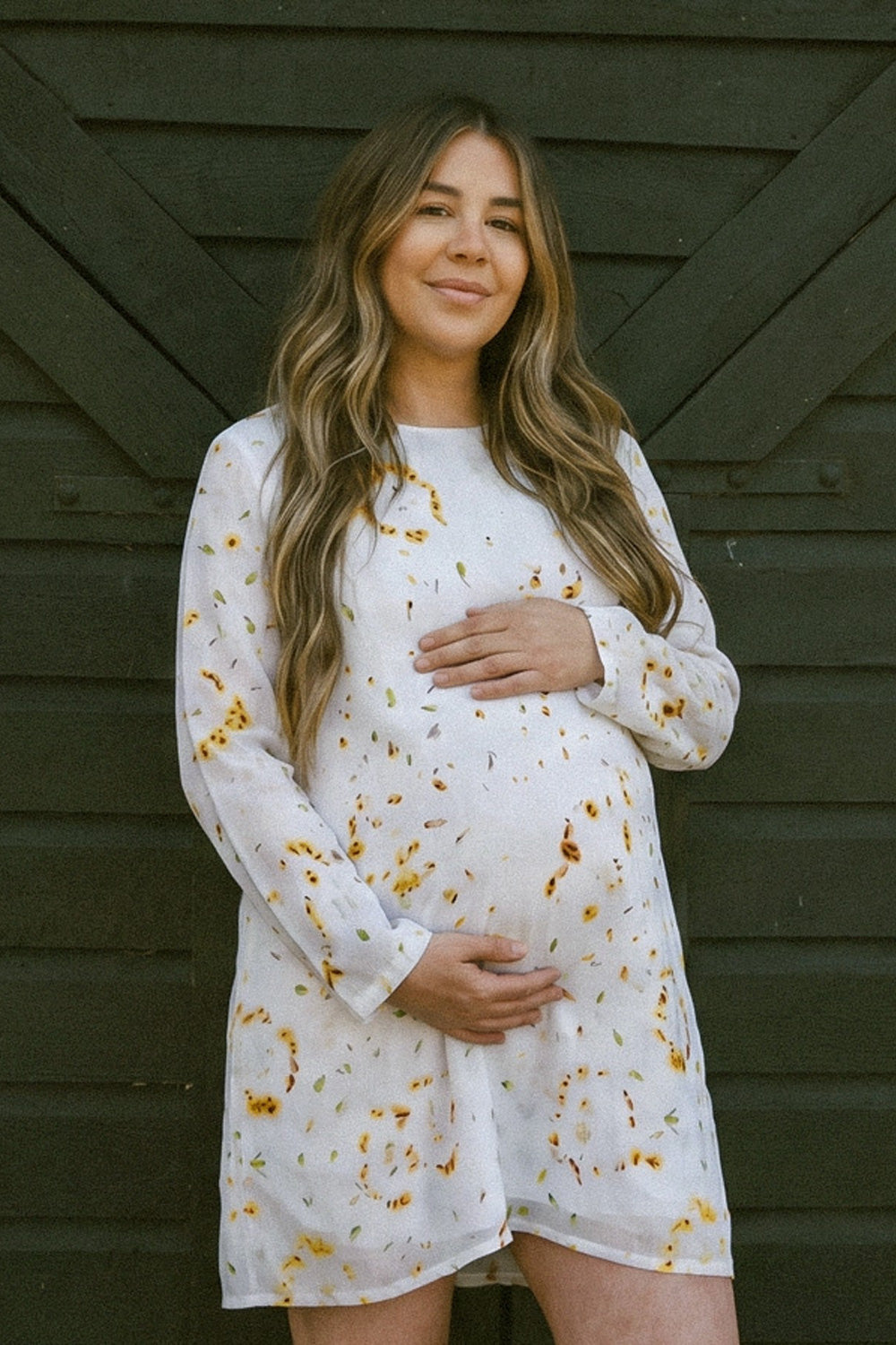 A pregnant woman wearing a white long-sleeve maternity linen dress with a yellow and orange eco-print, standing in front of a dark wooden wall.