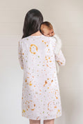 A woman seen from behind wearing a white long-sleeve linen dress with a delicate orange and green floral eco-print, while carrying a baby on her back. The background is a clean, off-white paneled wall.