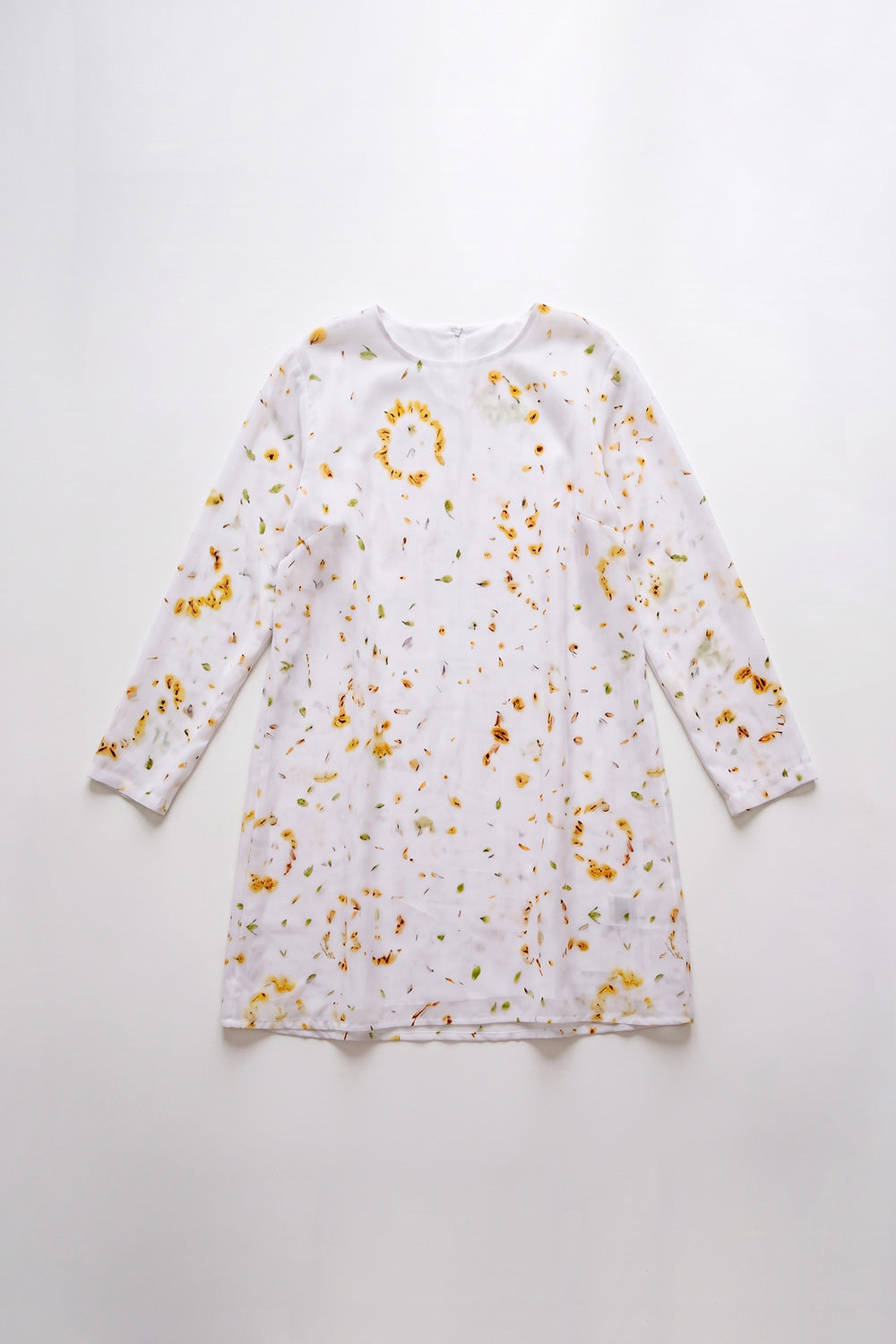A woman modeling a white long-sleeve shift linen dress with a yellow and orange eco-print, paired with white sneakers, against a light gray studio background.