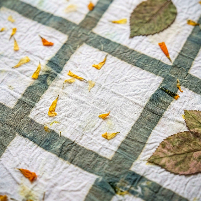 A close-up of a white white fabric with a muted green plaid pattern, scattered with dried orange flower petals and green leaves.