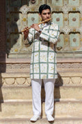 A man stands on stone steps in front of an ornate carved wall, holding a wooden flute. He is wearing a white kurta with a green grid pattern and small yellow floral motifs, paired with white trousers and white shoes.