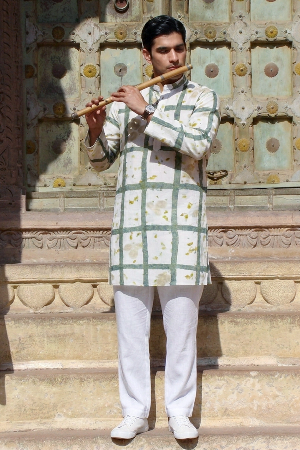 A man stands on stone steps in front of an ornate carved wall, holding a wooden flute. He is wearing a white kurta with a green grid pattern and small yellow floral motifs, paired with white trousers and white shoes.
