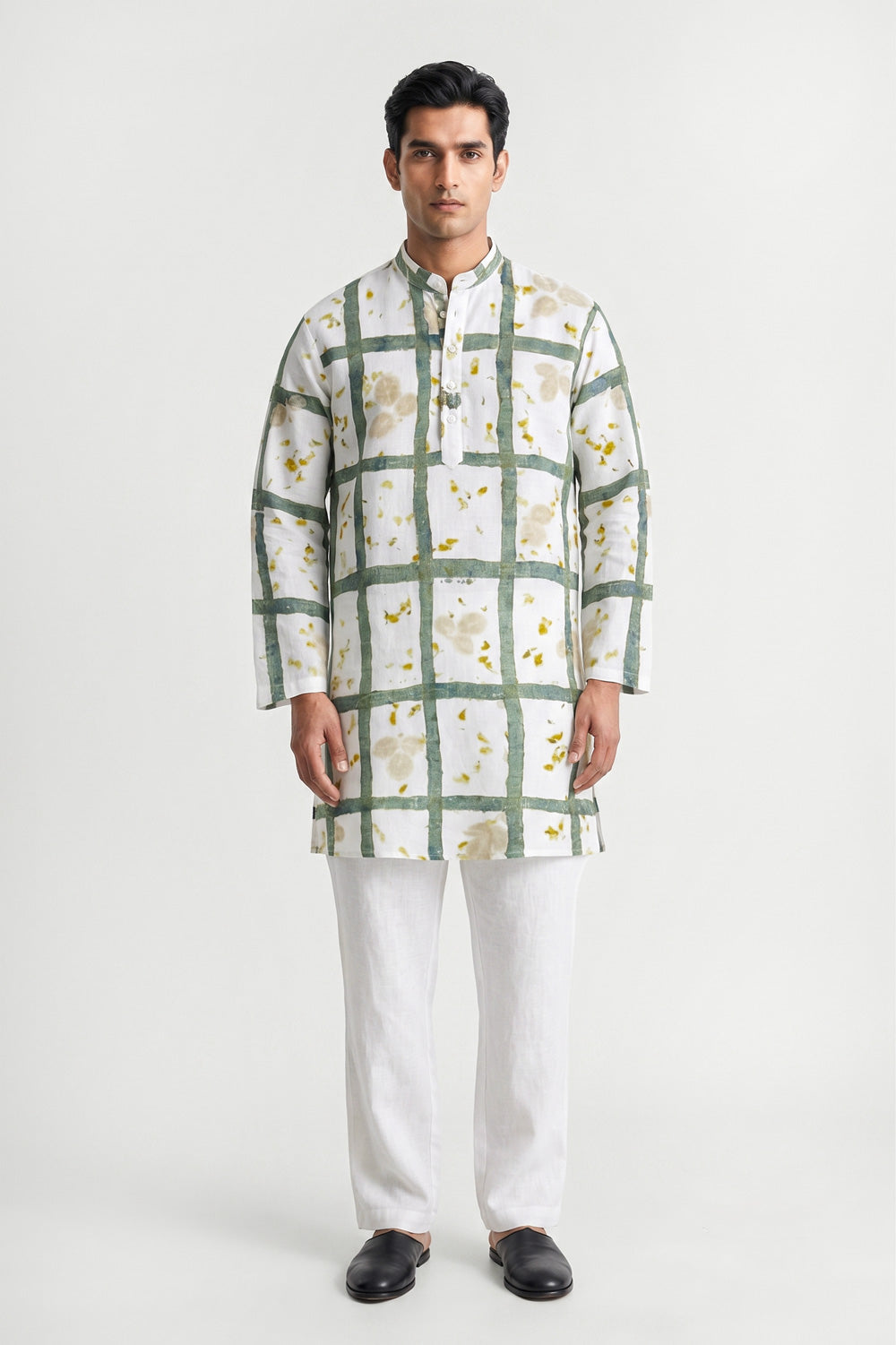 A man modeling a white long-sleeve kurta with a green grid pattern and small yellow floral accents, paired with white trousers and black shoes, against a light gray studio background.