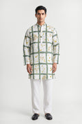 A man modeling a white long-sleeve kurta with a green grid pattern and small yellow floral accents, paired with white trousers and black shoes, against a light gray studio background.