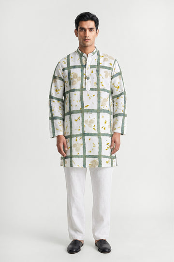 A man modeling a white long-sleeve kurta with a green grid pattern and small yellow floral accents, paired with white trousers and black shoes, against a light gray studio background.