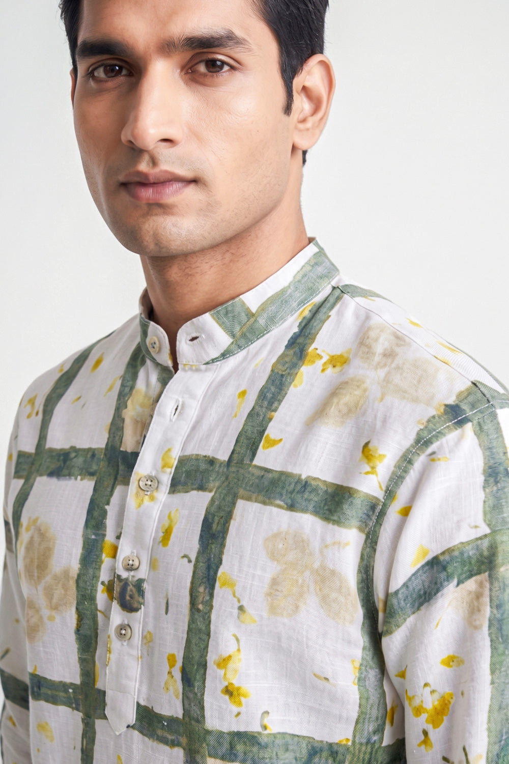A close-up of a man wearing a white kurta with a green grid pattern and yellow floral accents, set against a plain light gray background.