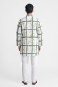 A man viewed from the back wearing a white long-sleeve kurta with a green grid pattern and small yellow floral details, paired with white trousers and black sandals, against a light gray studio background.