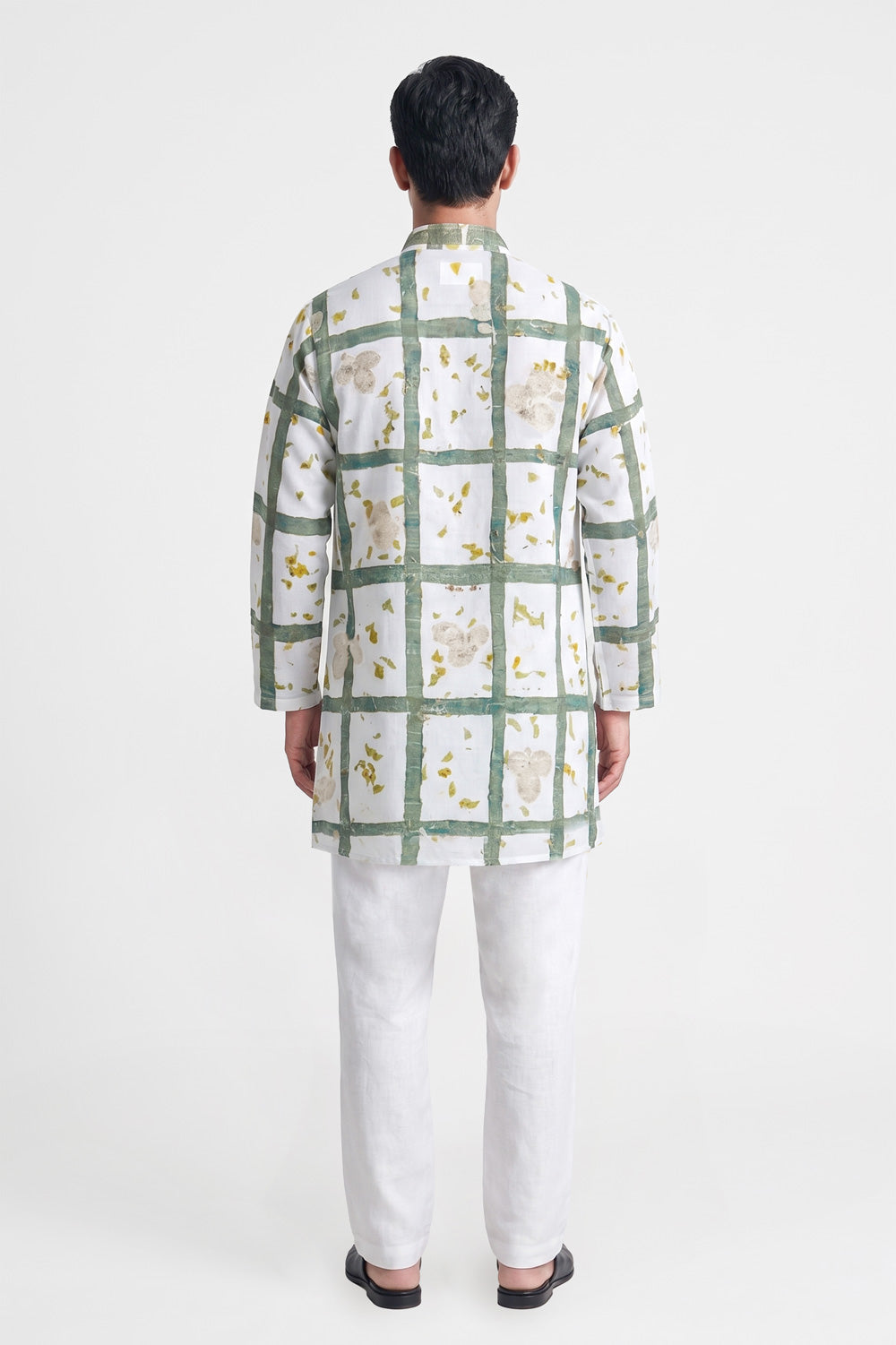 A man viewed from the back wearing a white long-sleeve kurta with a green grid pattern and small yellow floral details, paired with white trousers and black sandals, against a light gray studio background.