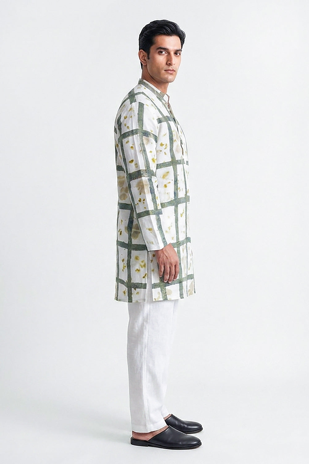A man wearing a white long-sleeve kurta with a green grid pattern and small yellow floral accents, paired with white trousers and black loafers, standing against a plain white studio background.