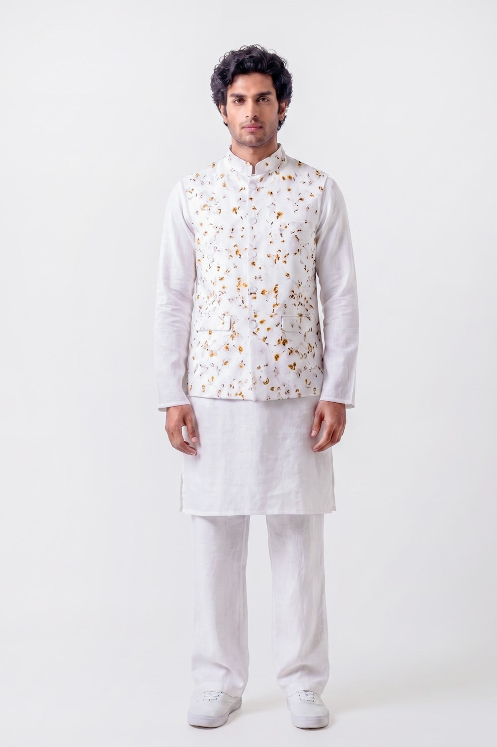 A man modeling a white traditional Indian outfit consisting of a kurta, trousers, and a white embroidered vest with gold and orange floral patterns, shot against a plain white studio background.