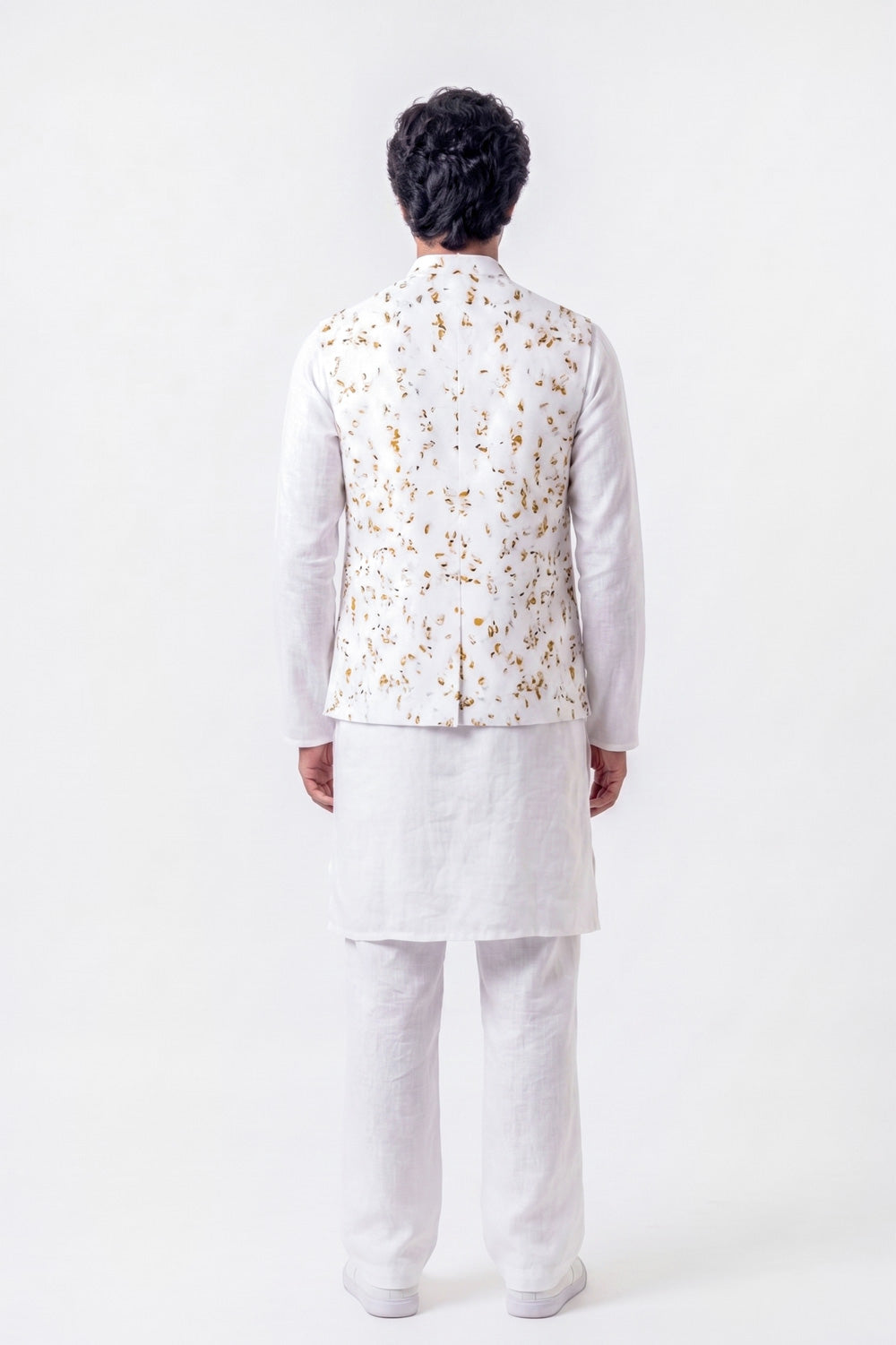 A man viewed from the back wearing a white traditional outfit with a white kurta, white trousers, and a white vest decorated with a gold floral pattern, set against a plain white background.
