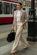 A woman walking on a city street wearing a striped linen blazer with pastel pink, blue, orange, and green stripes over a white top and cream wide-leg trousers, carrying a black tote bag.