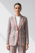A woman modeling a matching two-piece linen suit with vertical multicolor stripes in shades of blue, red, orange, yellow, green, and white, worn over a plain white top against a light gray studio background.