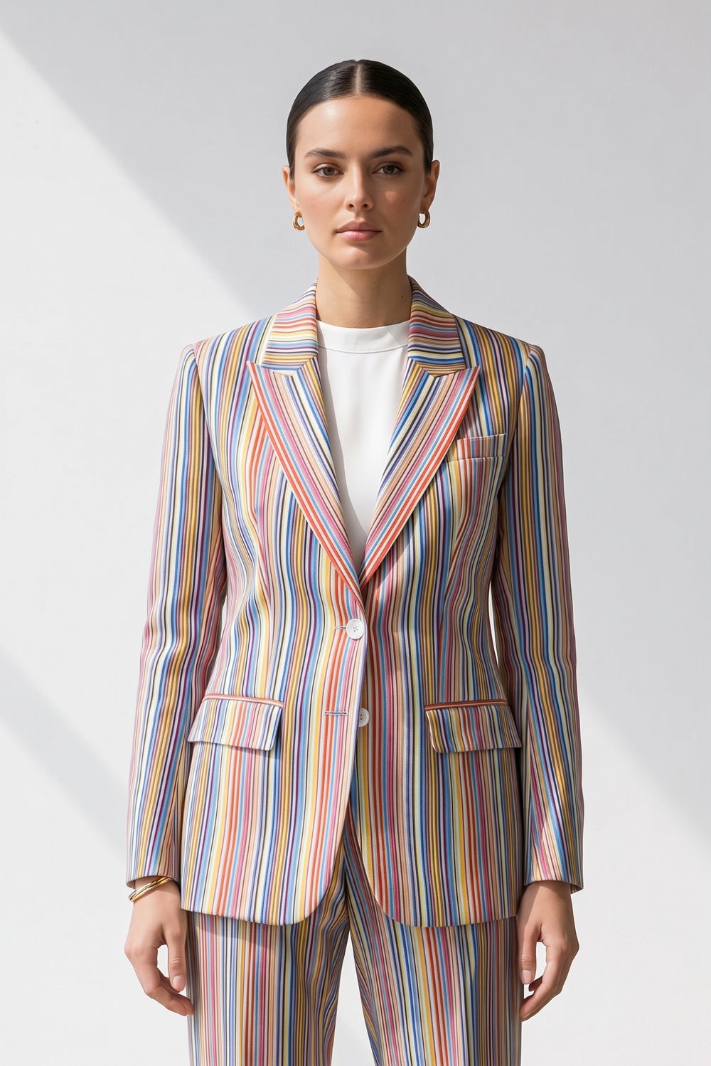 A woman modeling a matching two-piece linen suit with vertical multicolor stripes in shades of blue, red, orange, yellow, green, and white, worn over a plain white top against a light gray studio background.