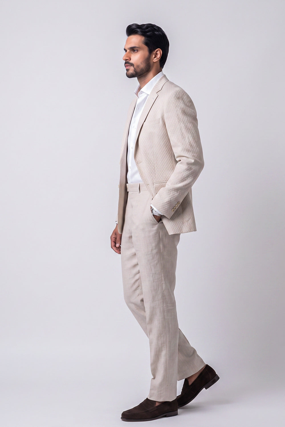 A man in a light beige textured linen suit with a white shirt and dark brown loafers, shown in profile against a light gray studio background.