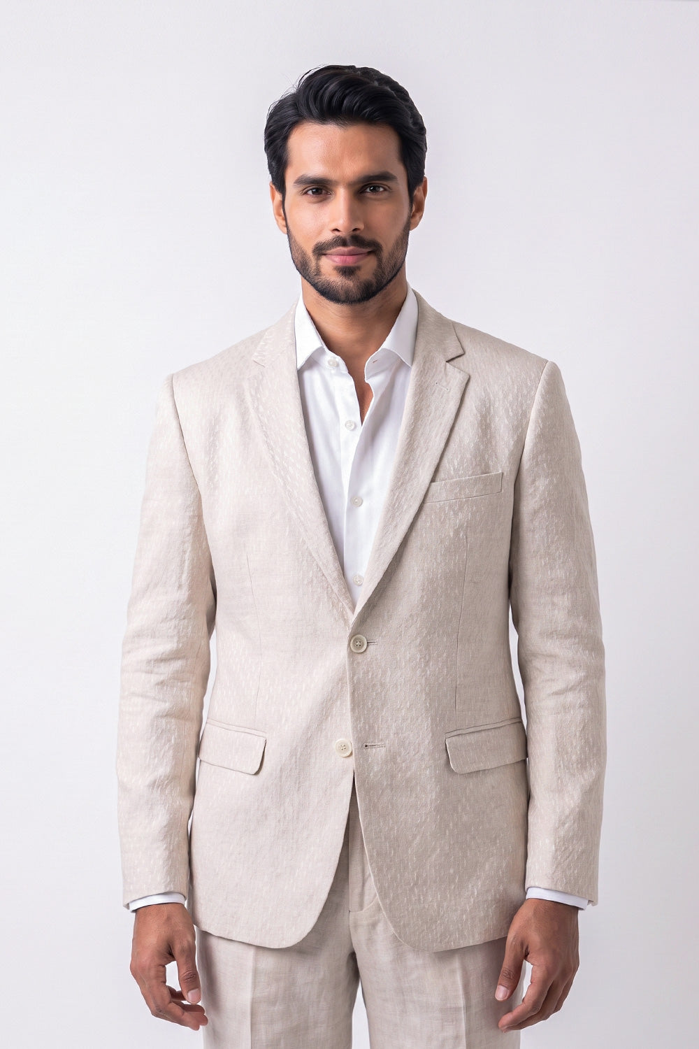 A man wearing a light beige textured linen blazer and matching trousers over a white dress shirt, standing against a plain light gray studio background.