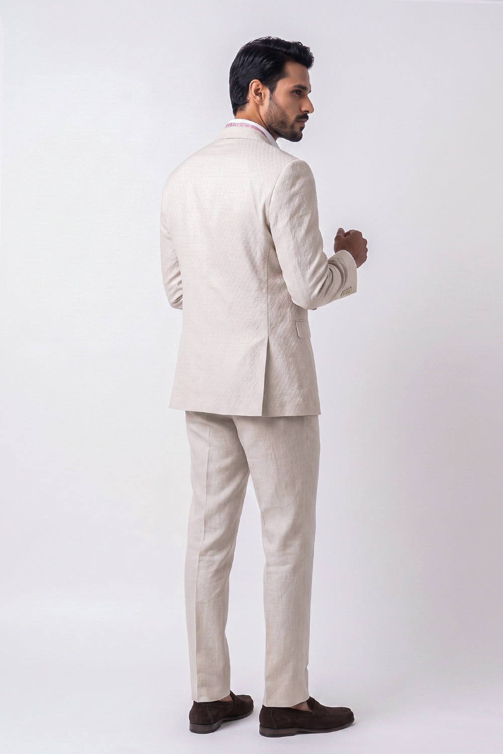 A man shown from the back wearing a light beige textured linen suit with a white shirt and dark brown loafers, standing against a plain white studio background.