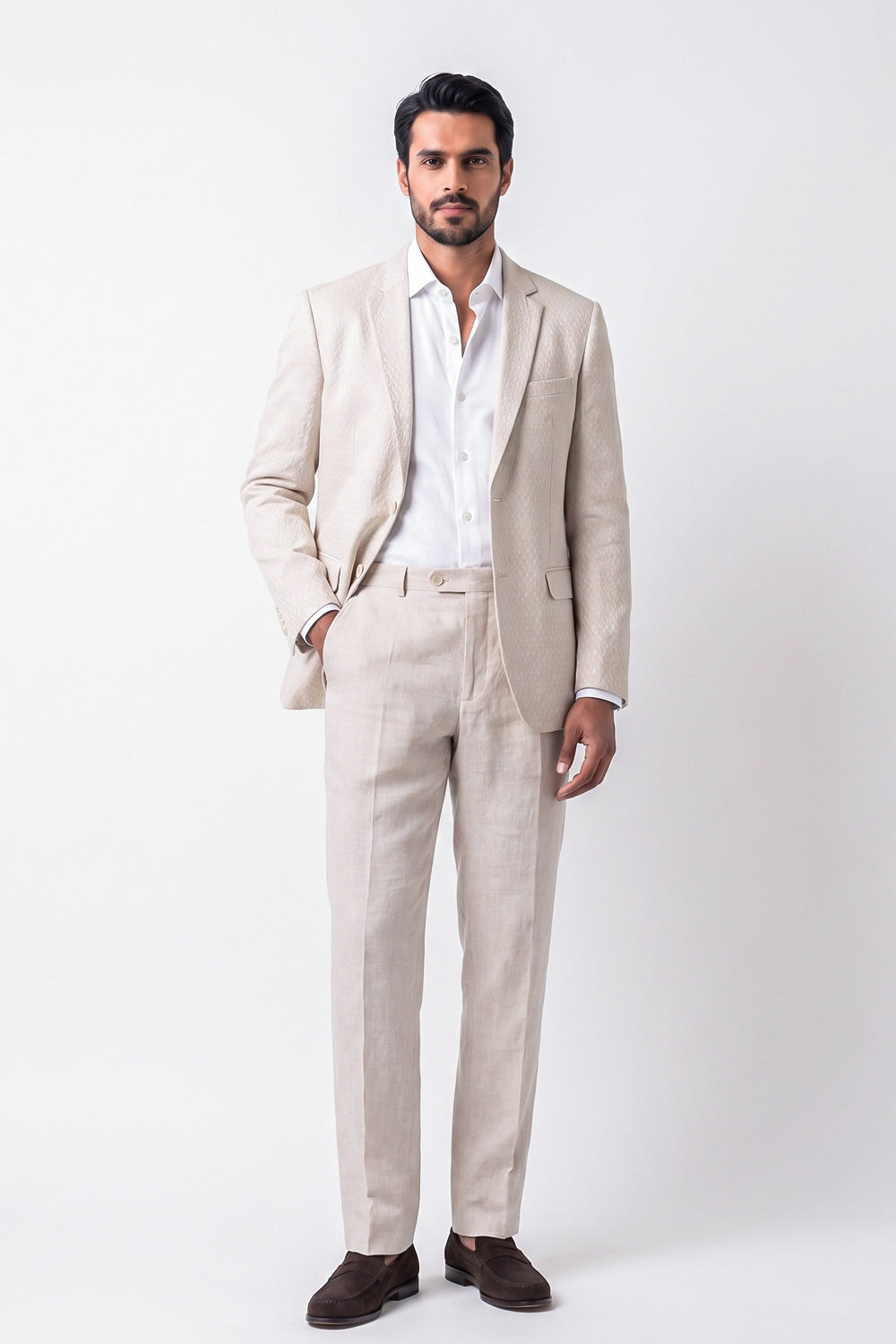 A man modeling a light beige linen suit with a white dress shirt and dark brown loafers, standing against a plain white studio background.