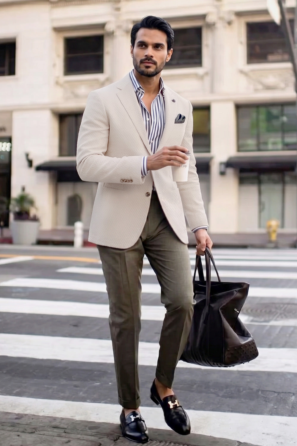 A man walking on a city crosswalk wearing a light beige linen blazer, olive green trousers, a blue and white striped shirt, and black loafers, while carrying a black tote bag.