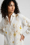 A woman with curly dark hair wearing a white button-up shirt with a yellow and brown watercolor floral print, photographed against a light gray studio background.
