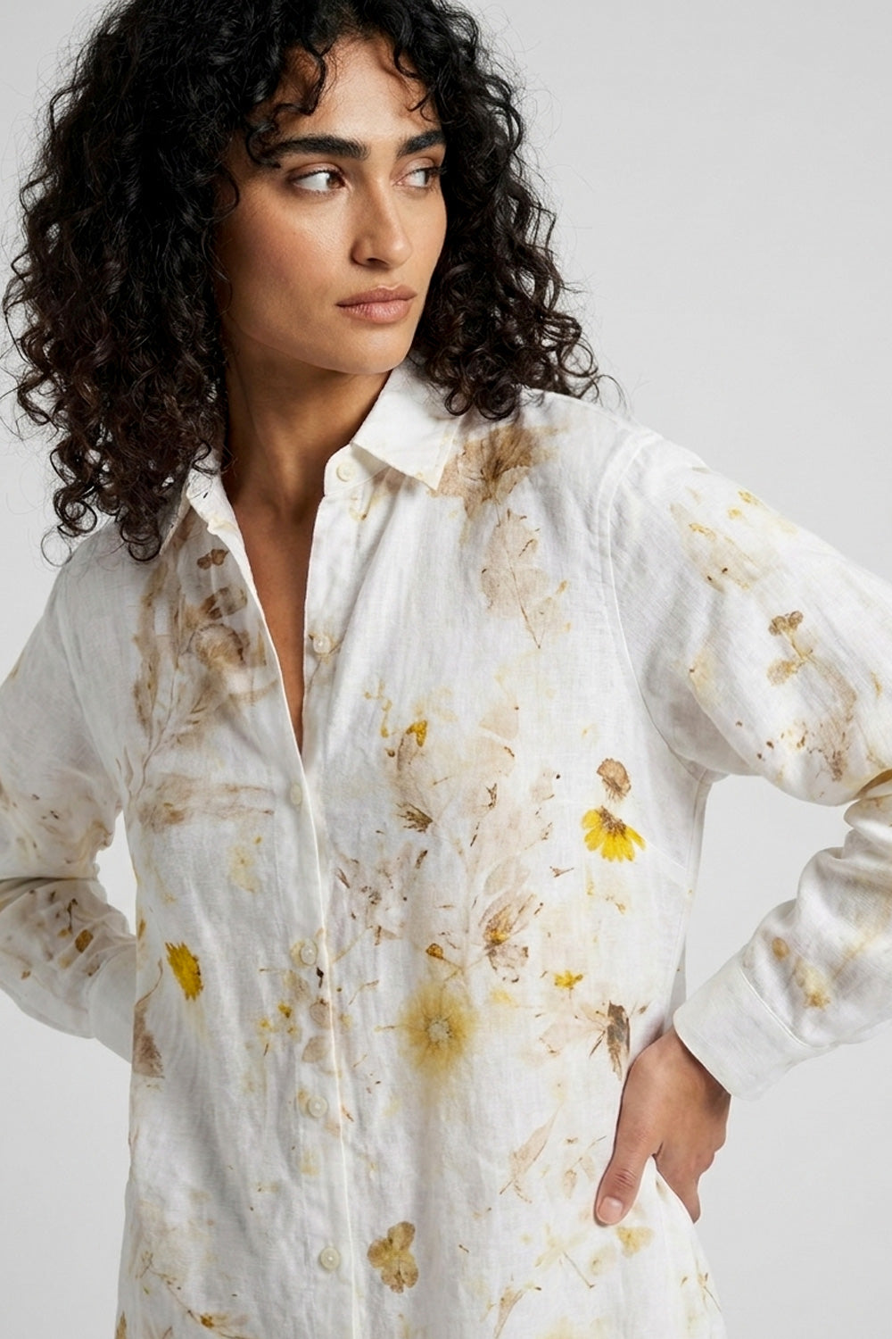 A woman with curly dark hair wearing a white button-up shirt with a yellow and brown watercolor floral print, photographed against a light gray studio background.
