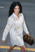 A woman walking on a street wearing a white long-sleeve shirt dress with yellow and brown botanical prints splatters, holding a brown Louis Vuitton monogram duffel bag.