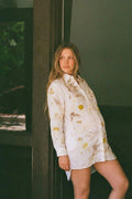 A pregnant woman leans against a dark pillar wearing a white long-sleeve  shirt dress with a yellow and brown botanical print.