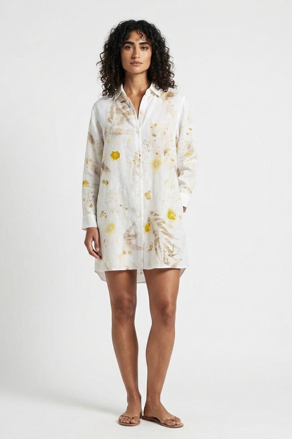 A woman modeling a white long-sleeve button-up shirt dress with a yellow and beige botanical print, standing against a plain white studio background.