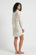 A woman modeling a long-sleeve white shirt dress with a yellow and gold abstract floral pattern, shown from a side-back angle against a plain white studio background.